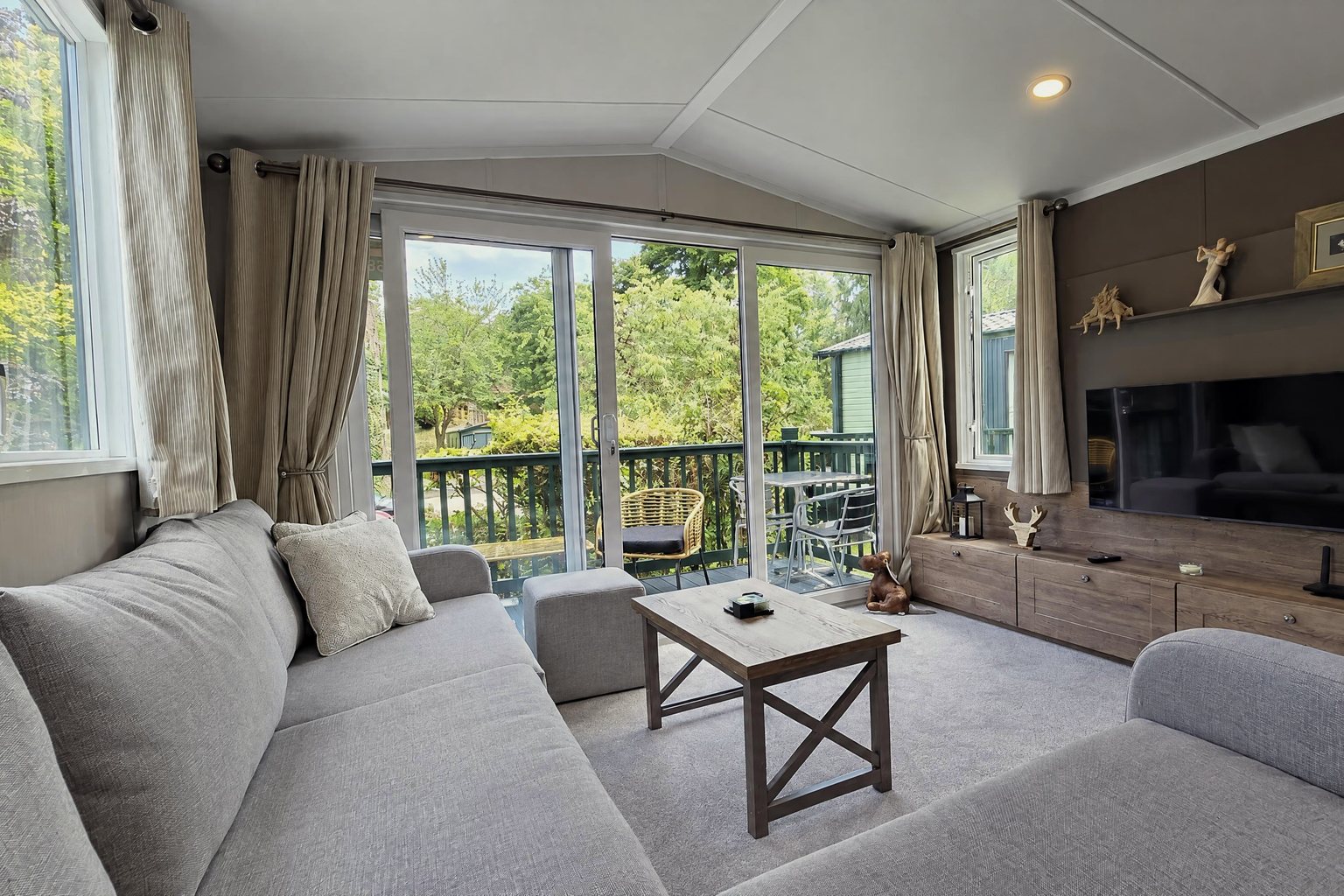 The living room in daylight: corner sofa, coffee table, TV wall and glass doors onto the deck