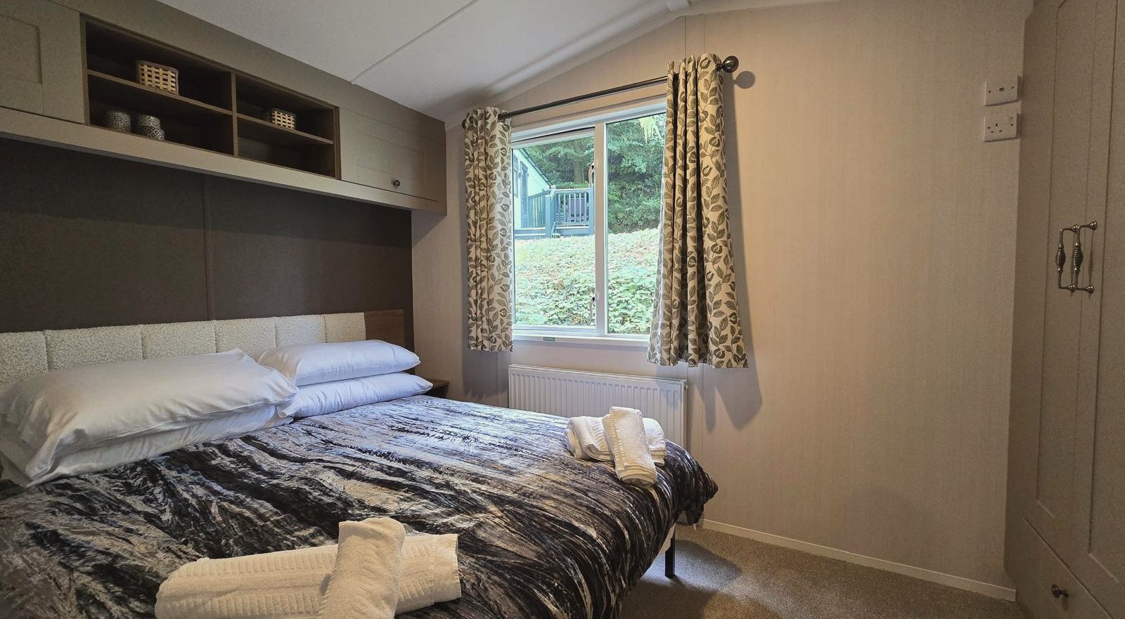 A double bedroom with a padded headboard, shelving above the bed and patterned curtains
