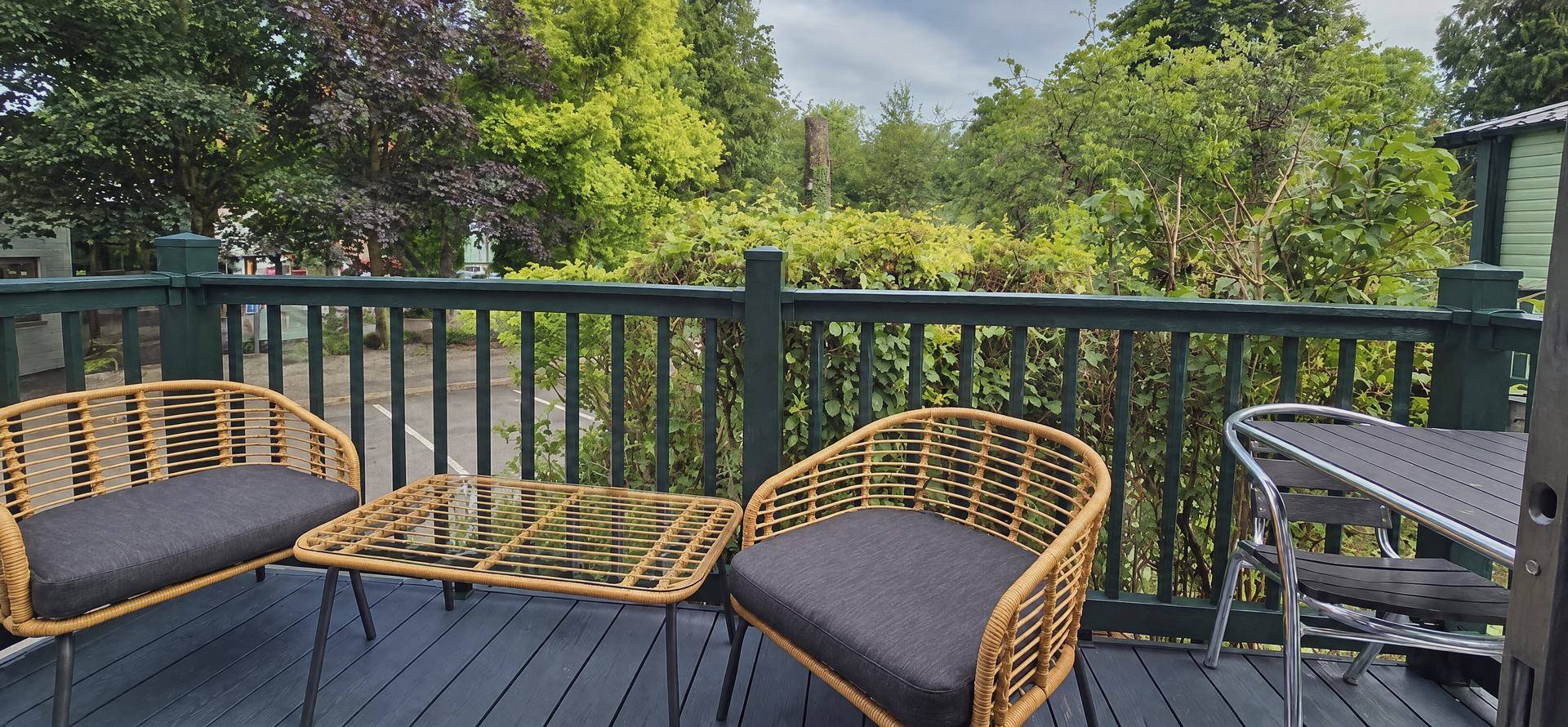 A deck with a pair of rattan armchairs, a glass-topped table and a metal bistro set, trees beyond the rails