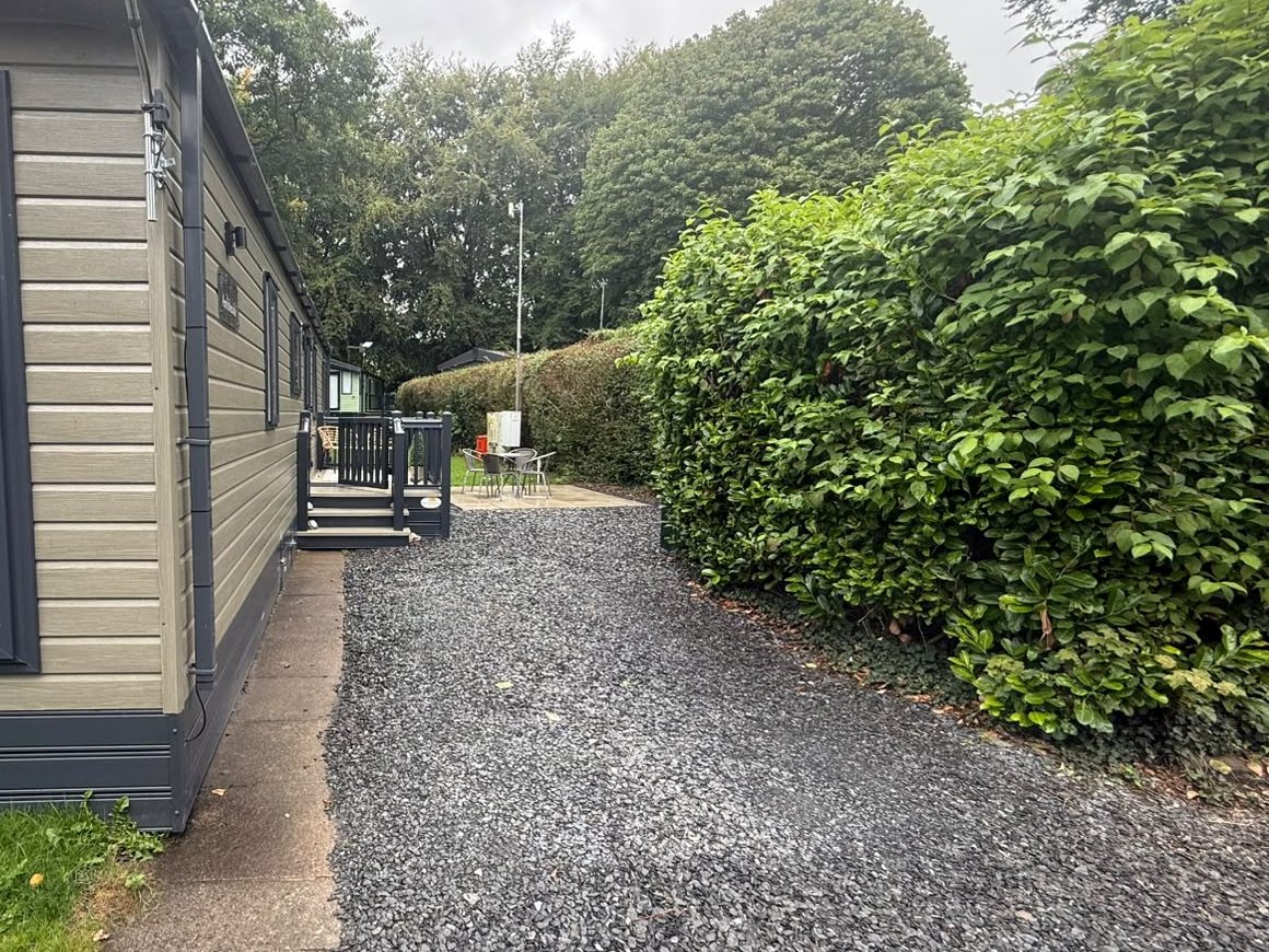 The side of the holiday home along a gravel path, with a tall hedge opposite