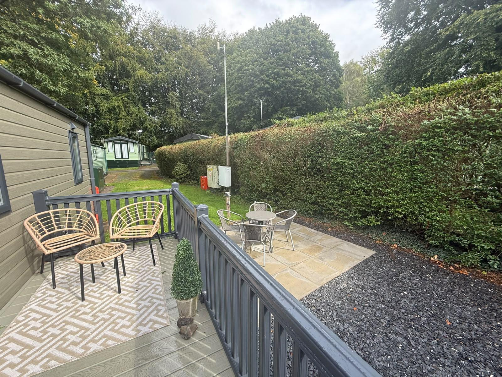 The deck and the paved patio together: two rattan chairs and a low table on a patterned rug, a bistro table and four chairs on the patio beyond the rails, a tall hedge alongside